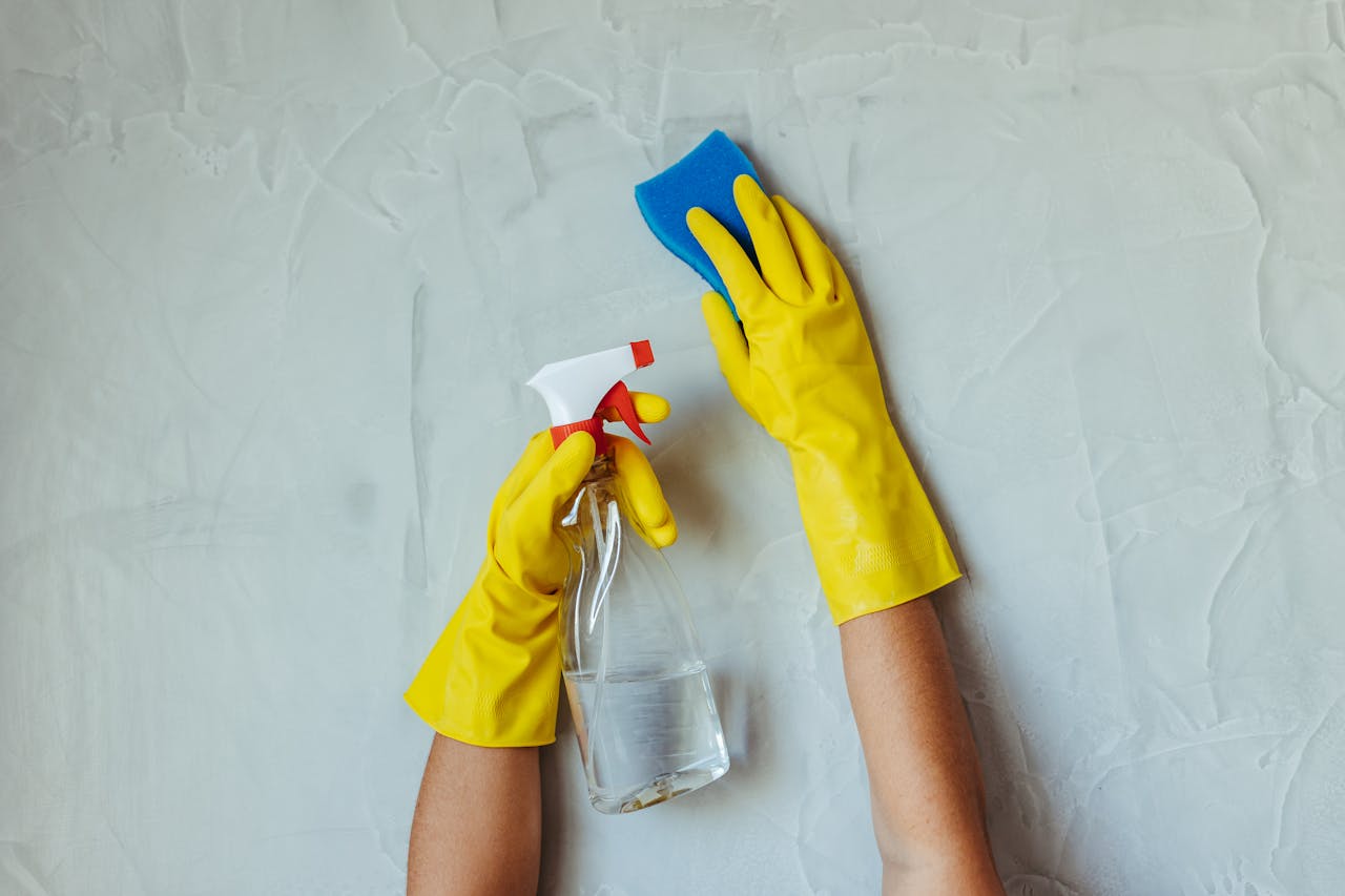 Entreprise de nettoyage à Dijon, Ahuy et à Bressey-sur-Tille avec SOS DC Close-up of hands in yellow gloves cleaning a wall with a spray bottle and sponge.