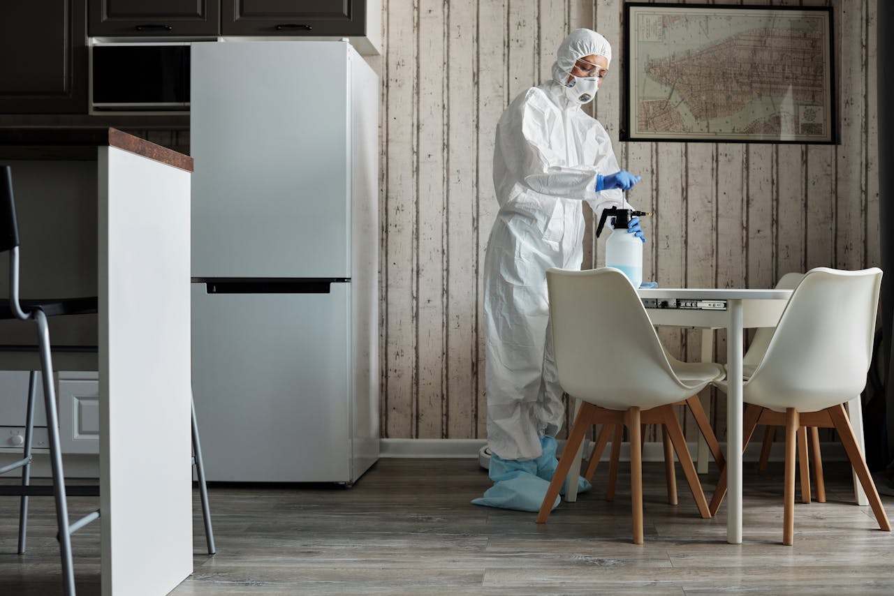 Les tendances de nettoyage extrême à tester cette année Person in hazmat suit disinfecting a modern kitchen table with spray bottle for hygiene.